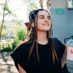 Smiling young woman with a bandana and braids holds a beverage while enjoying a sunny day outside.